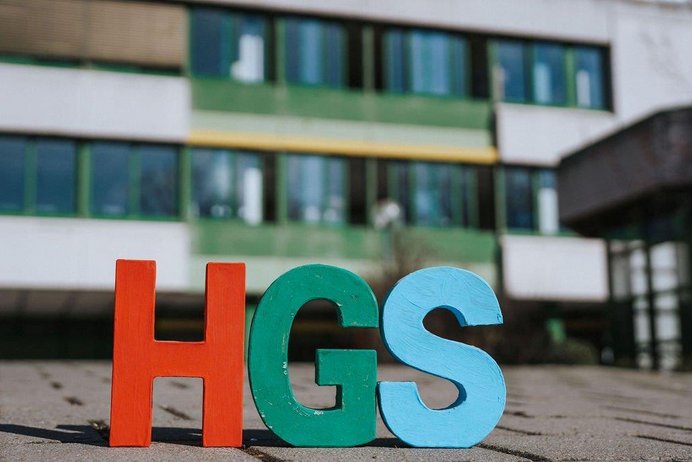Große Buchstaben HGS vor dem unscharfen Schulgebäude im Hintergrund. 